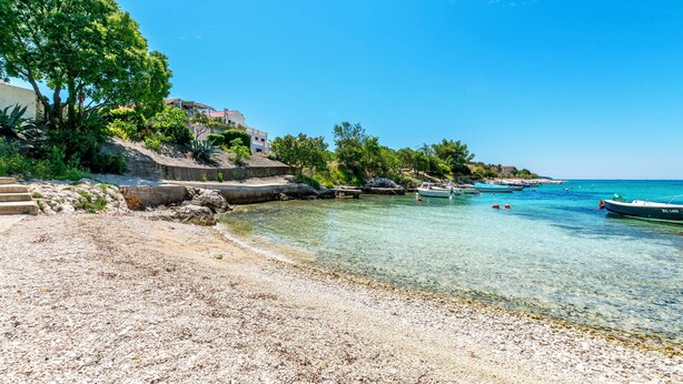 Μια παραθαλάσσια παραλία με καθαρά νερά, βάρκες και ένα κτίριο με σκαλοπάτια που οδηγούν προς την ακτή.
