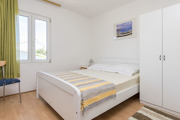 A bedroom with a bed, a nightstand, a chair, a window with curtains, a wardrobe, and a painting on the wall. - Apartments Ljubomir (Rogoznica)