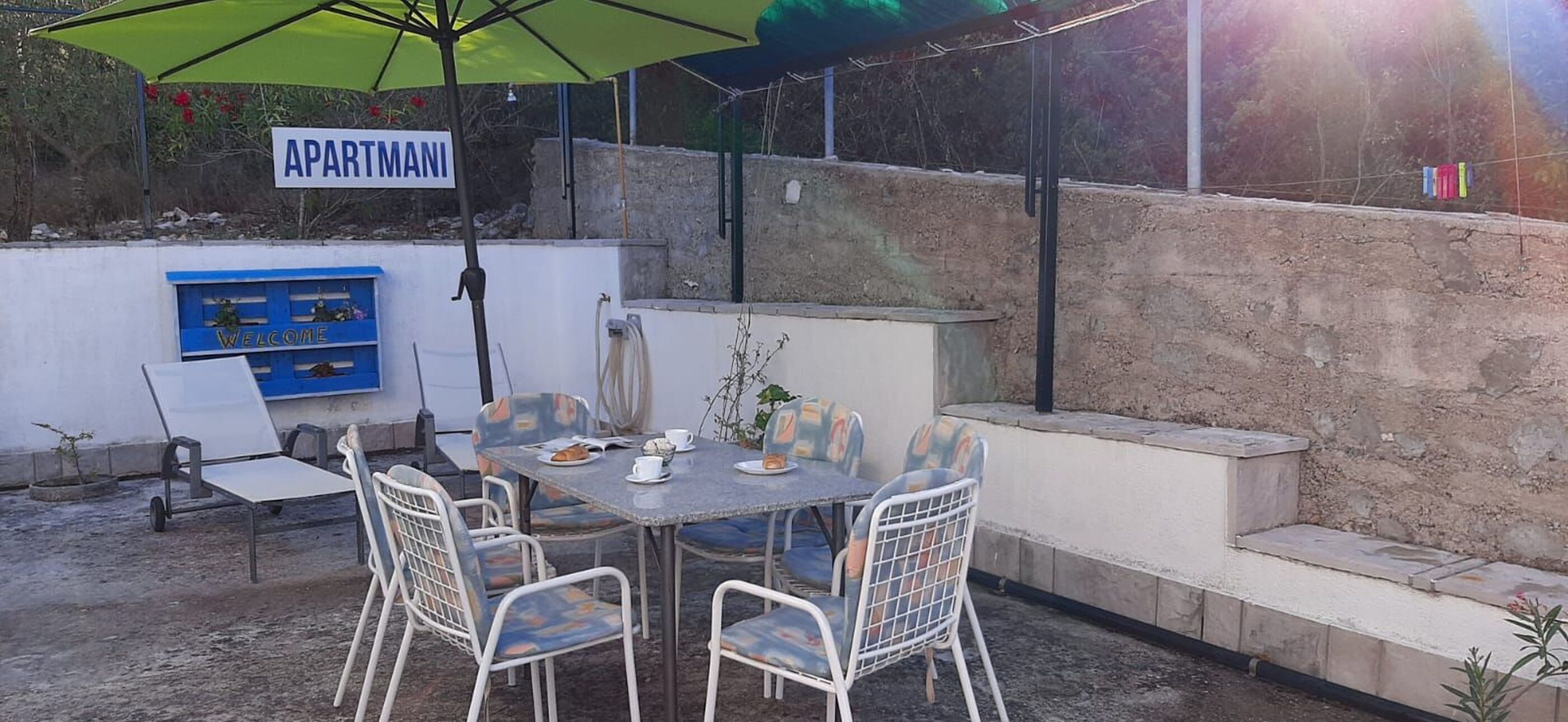 An outdoor patio with a table set for two, surrounded by chairs, under a green umbrella. A sign reading "APARTMANI" is visible in the background.