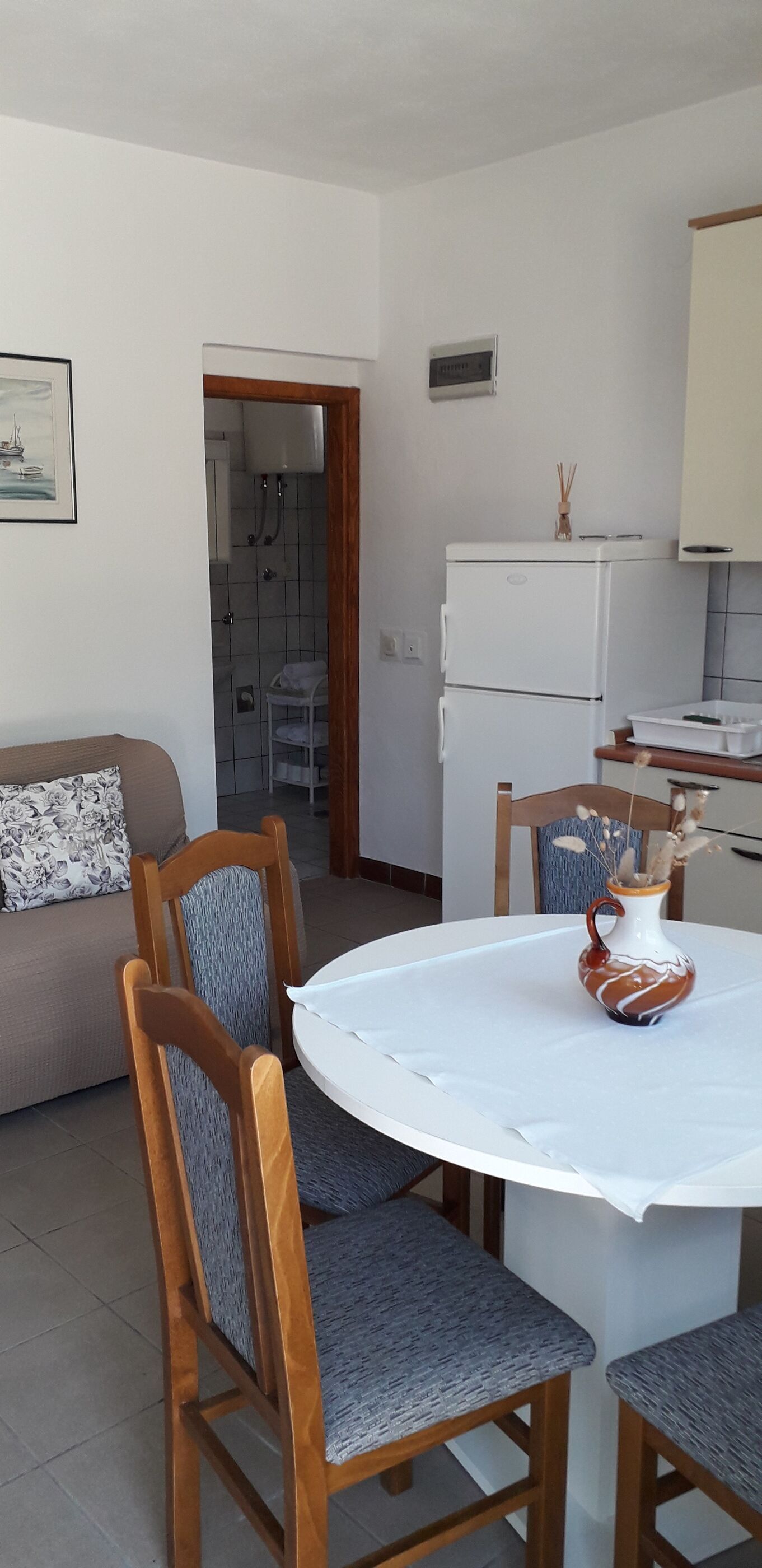 Un ambiente compatto con zona pranzo con tavolo rotondo, sedie in legno e tovaglia bianca. Si vedono anche un frigorifero e un piccolo mobiletto nella zona cucina.