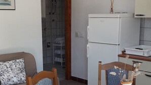 Ein kompakter Wohnbereich mit Essplatz, der einen runden Tisch, Holzstühle und eine weiße Tischdecke umfasst. Im Küchenbereich sind ein Kühlschrank und eine kleine Vitrine erkennbar.