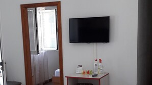 Ein Wohnzimmer mit einer Couch, einem an der Wand montierten Fernseher, einem kleinen Tisch mit Gegenständen und einer Tür zu einem anderen Raum.