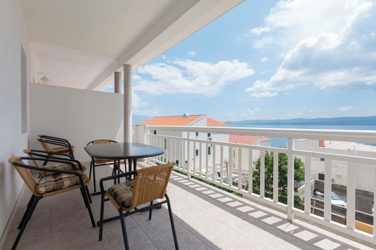 A balcony with a table and chairs, a white railing, and a view of buildings and mountains in the distance.