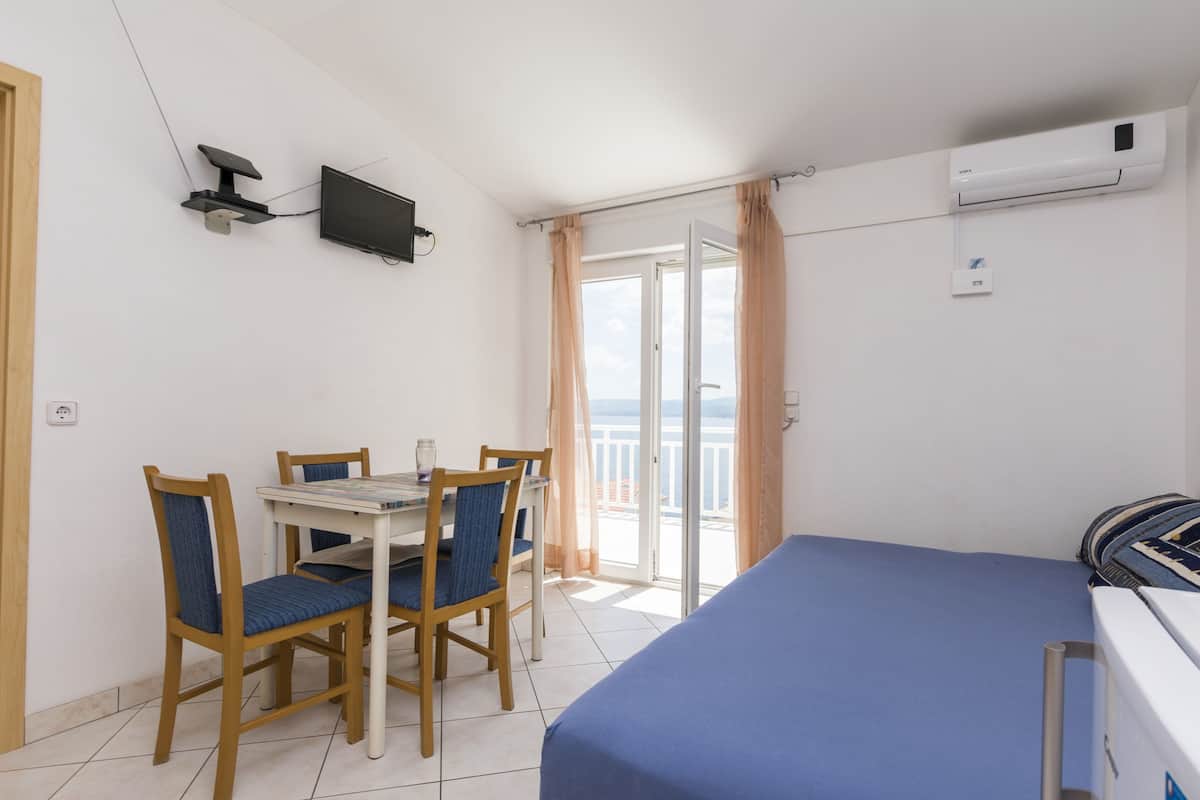 A room with a bed, a TV mounted on the wall, a dining table with chairs, and a view of the sea through a glass door.