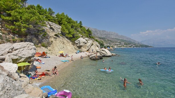 Ein Strand mit klarem Wasser, felsigem Gelände und Menschen, die schwimmen und sich sonnen.
