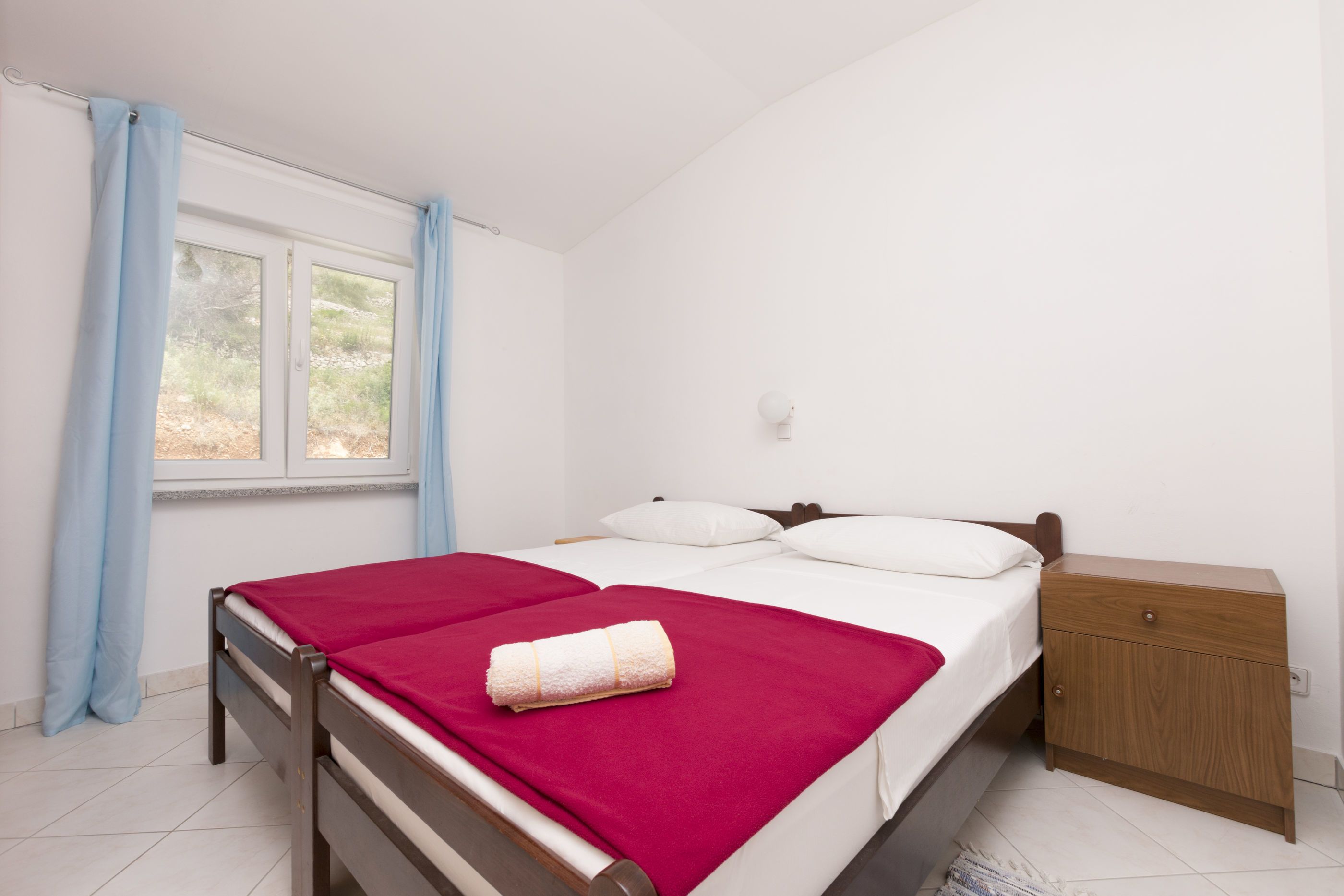 A bedroom with a bed, a nightstand, a window with curtains, and a view of the outdoors.
