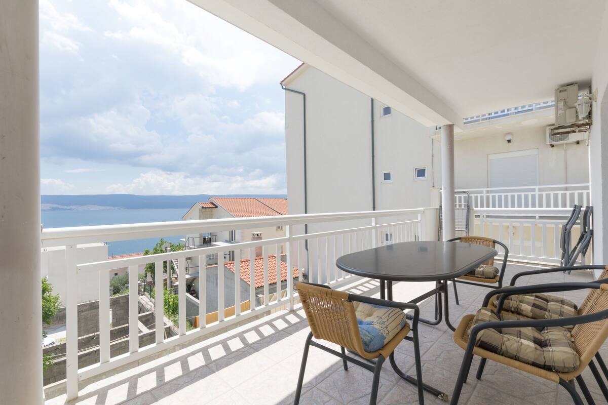 A balcony with a table and chairs, a white railing, and a view of the sea and buildings.