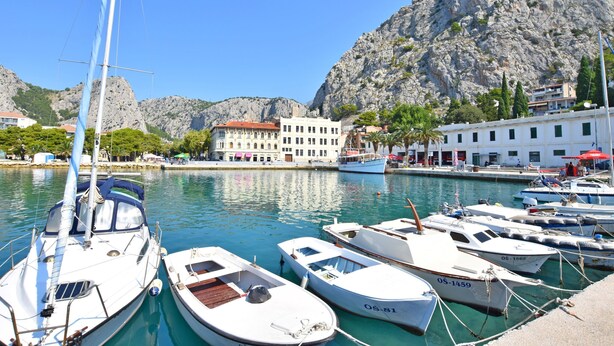 Un port de plaisance où sont amarrés plusieurs bateaux, un bâtiment avec un toit rouge et, au loin, des collines rocheuses.