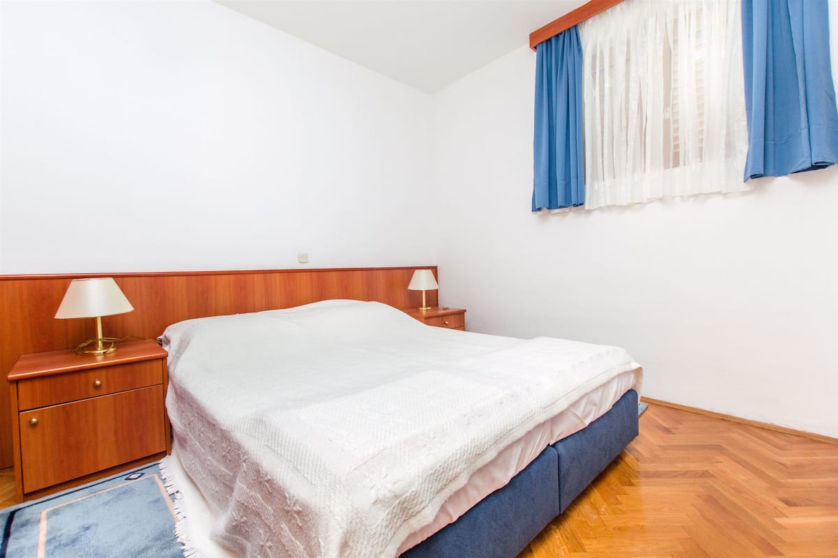 A bedroom with a wooden headboard, a single bed with a white blanket, a nightstand with a lamp, and a window with blue and white curtains.