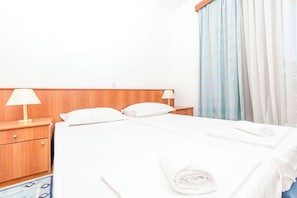Hotellihuone, jossa on puinen sängynpääty, kaksi yöpöytää valaisimineen, valkoisilla lakanoiden peitetyt sänky ja sängyllä rullalle taitettu pyyhe.