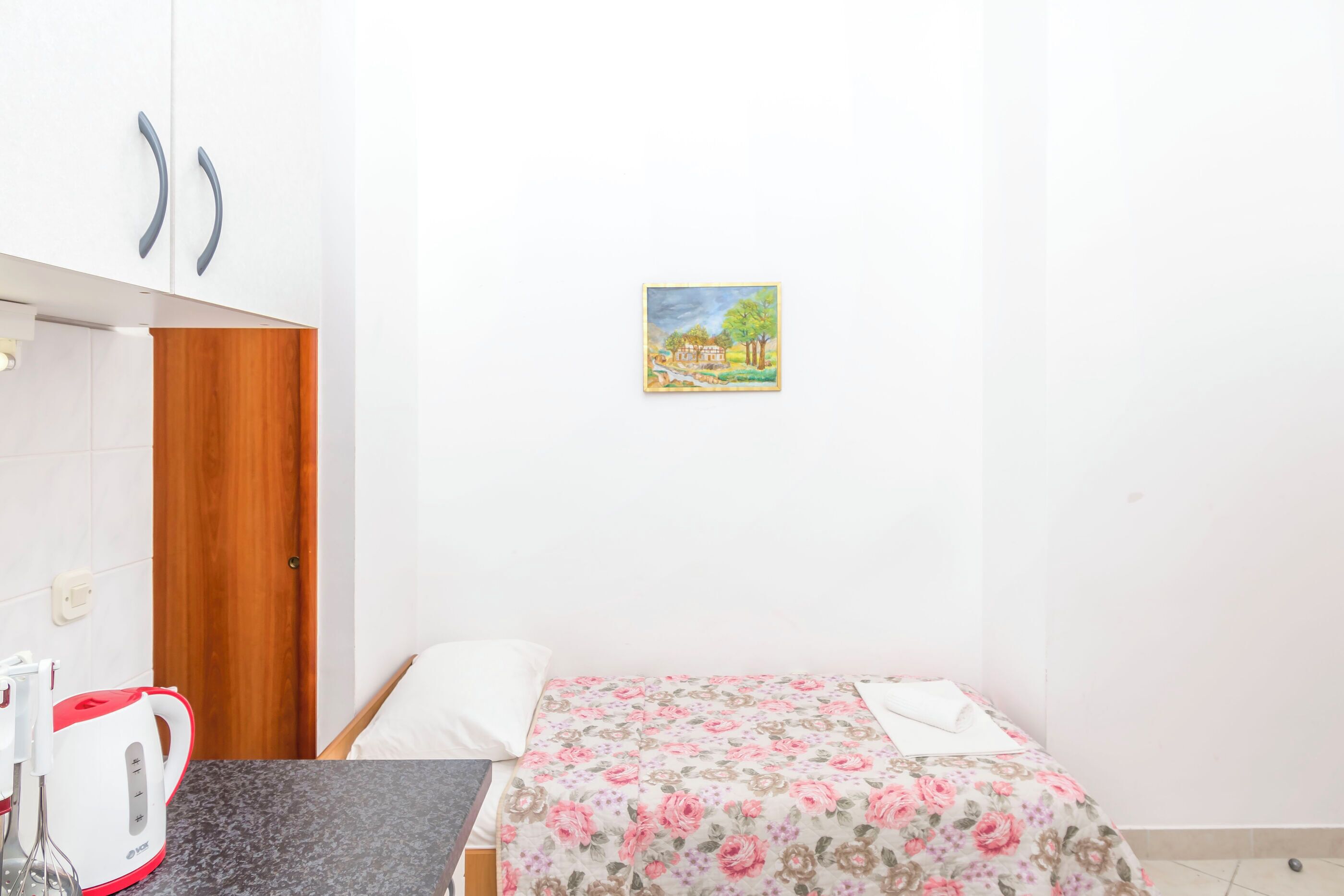 A small bedroom with a bed, a kettle, a wooden wardrobe, and a painting on the wall.