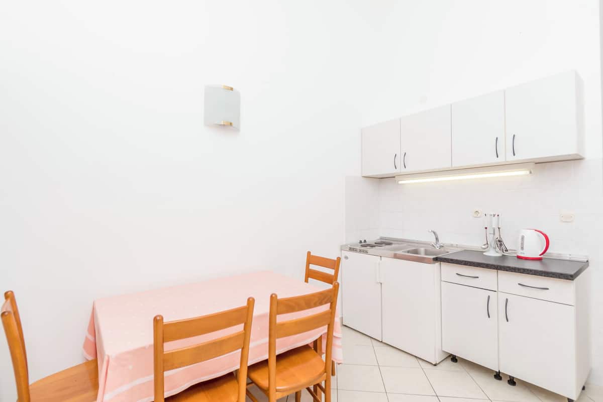 A small kitchenette with white cabinets, a sink, and a countertop. There is a table with chairs and a light fixture above the cabinets.
