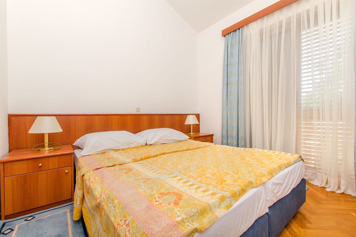 A bedroom with a wooden headboard, a bed with a patterned bedspread, two bedside lamps, and a window with sheer curtains.