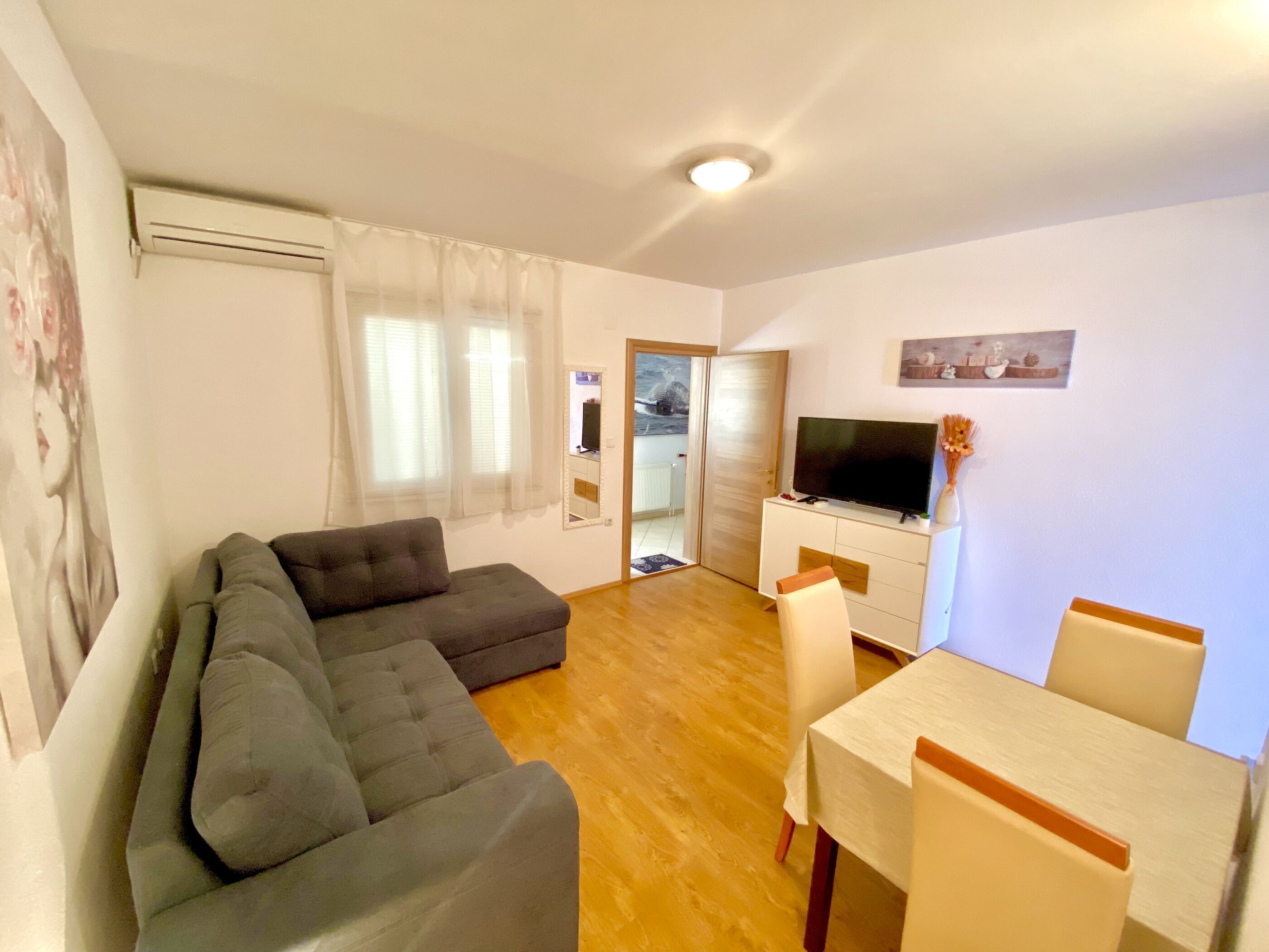 Een woonkamer met een grijze bank, een eettafel met stoelen, een televisie op een witte kast, en een raam met dunne gordijnen.