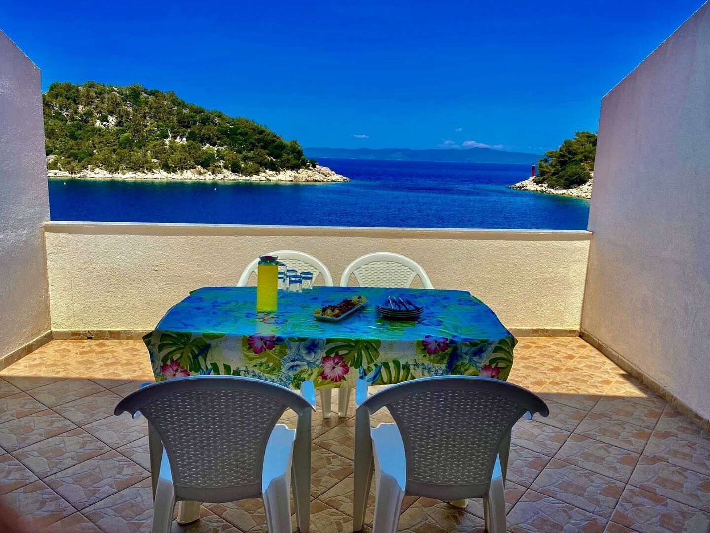 A table with two chairs on a balcony overlooking a beach with clear blue waters and a distant shoreline.