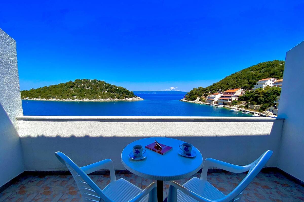 A balcony with a table and two chairs overlooking a coastal view with buildings and greenery.