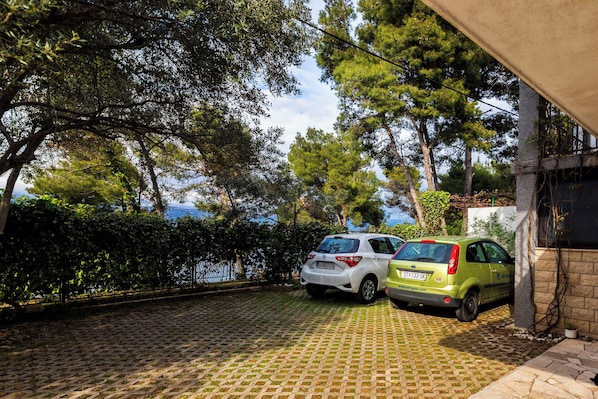 A paved driveway with two cars parked, surrounded by trees and a body of water in the background.