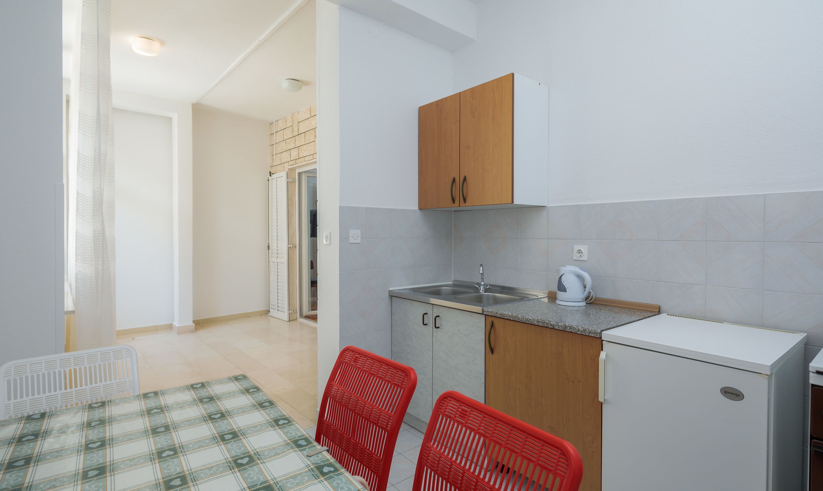 A compact kitchen with a sink, a kettle, and a refrigerator. There are two red chairs and a table with a checkered tablecloth.