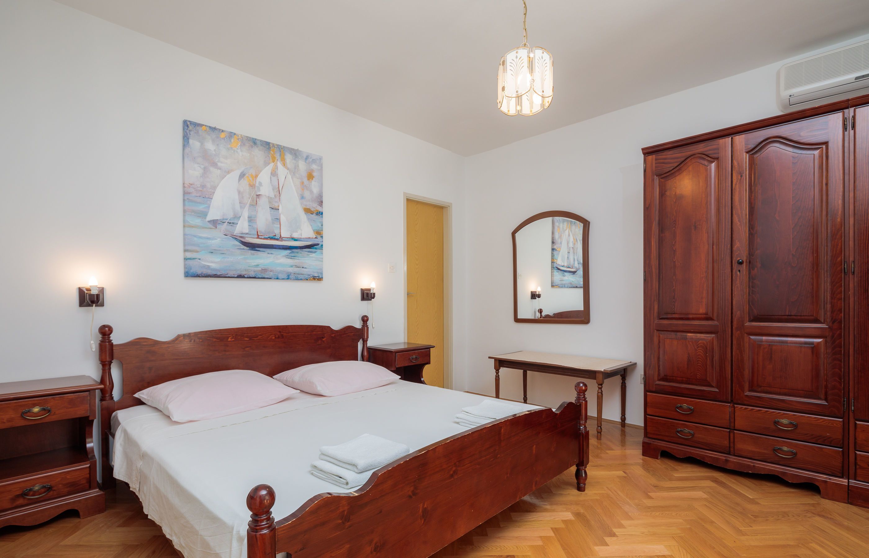 A bedroom with a wooden bed, bedside tables, a dresser, a painting of a sailboat, and a mirror.