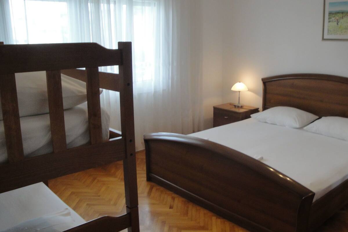 Una cama de madera con ropa de cama blanca en una habitación con piso de madera y una mesita de noche con una lámpara.