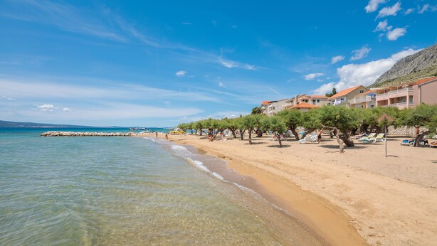 A beach with clear waters, a rocky breakwater, and buildings with balconies overlooking the sea. - Apartments and Rooms Denis (Duce)