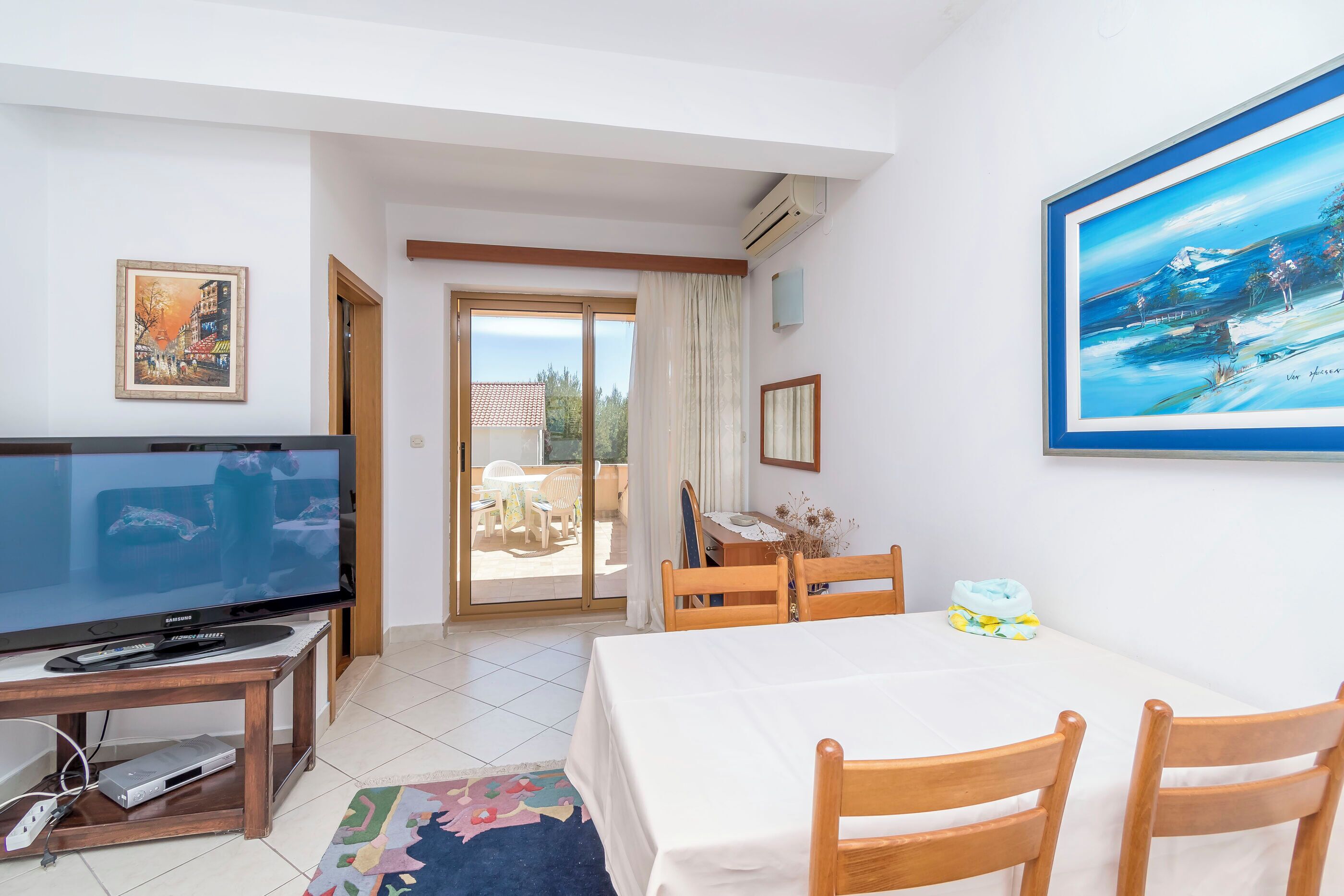 A room with a TV, a dining table with chairs, a painting, and a view through a glass door.