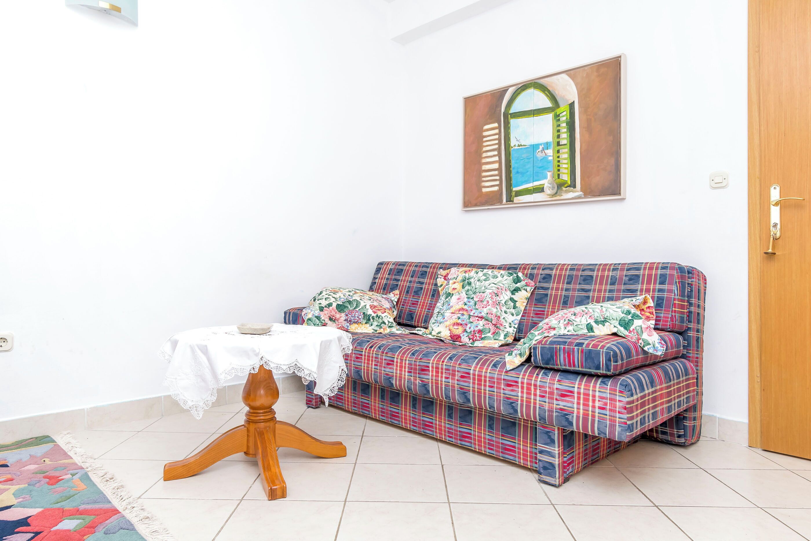 Une pièce comprenant un canapé, une petite table recouverte d’une nappe en dentelle, une chaise en bois et une porte.