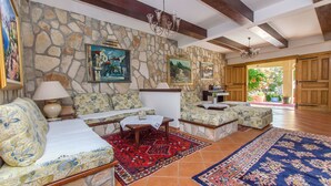 A cozy living room with stone walls, wooden beams, and a large window. There are two sofas, a coffee table, and a rug.