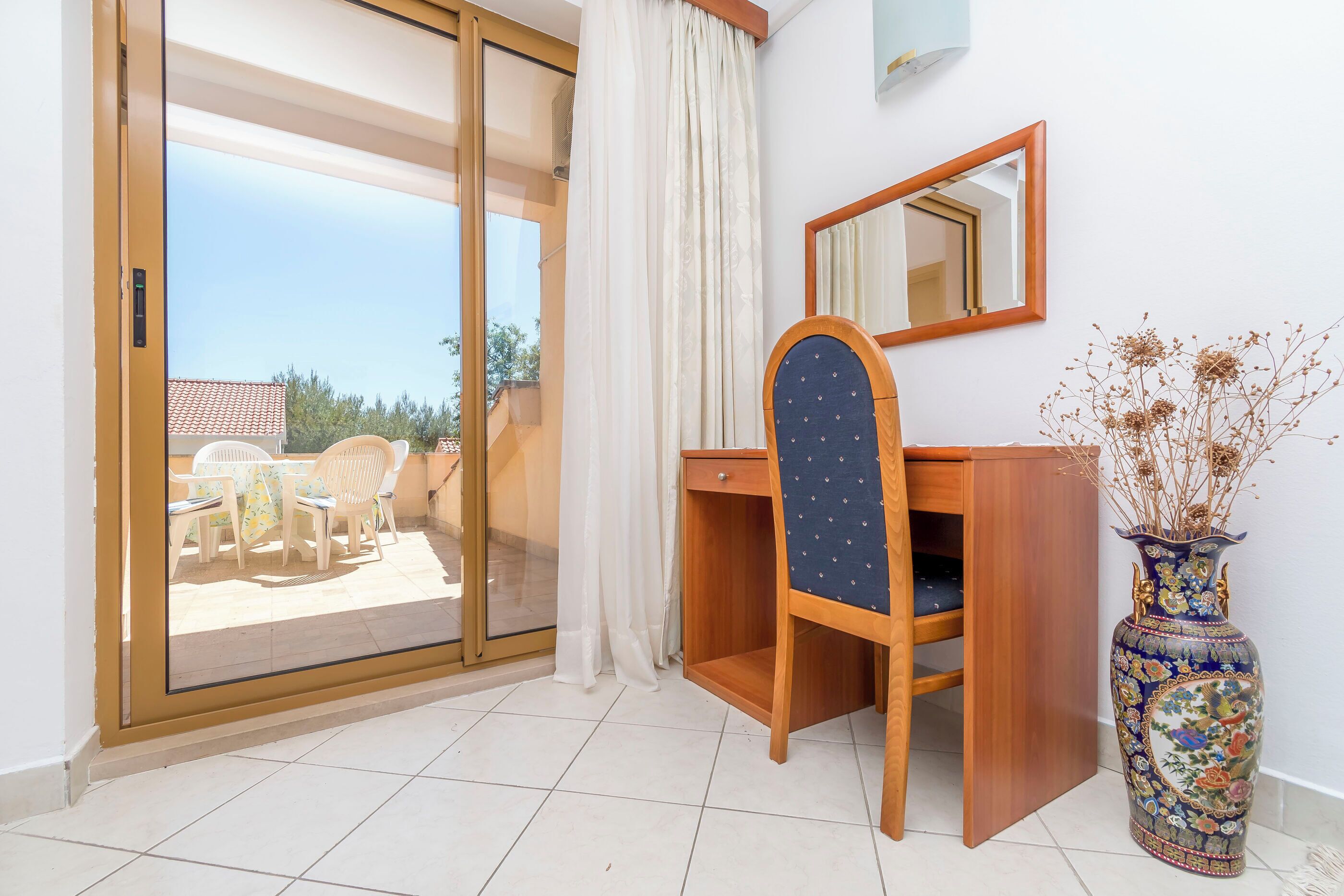 A room with a sliding glass door leading to an outdoor patio, a wooden desk, a chair, a mirror, and a decorative vase.