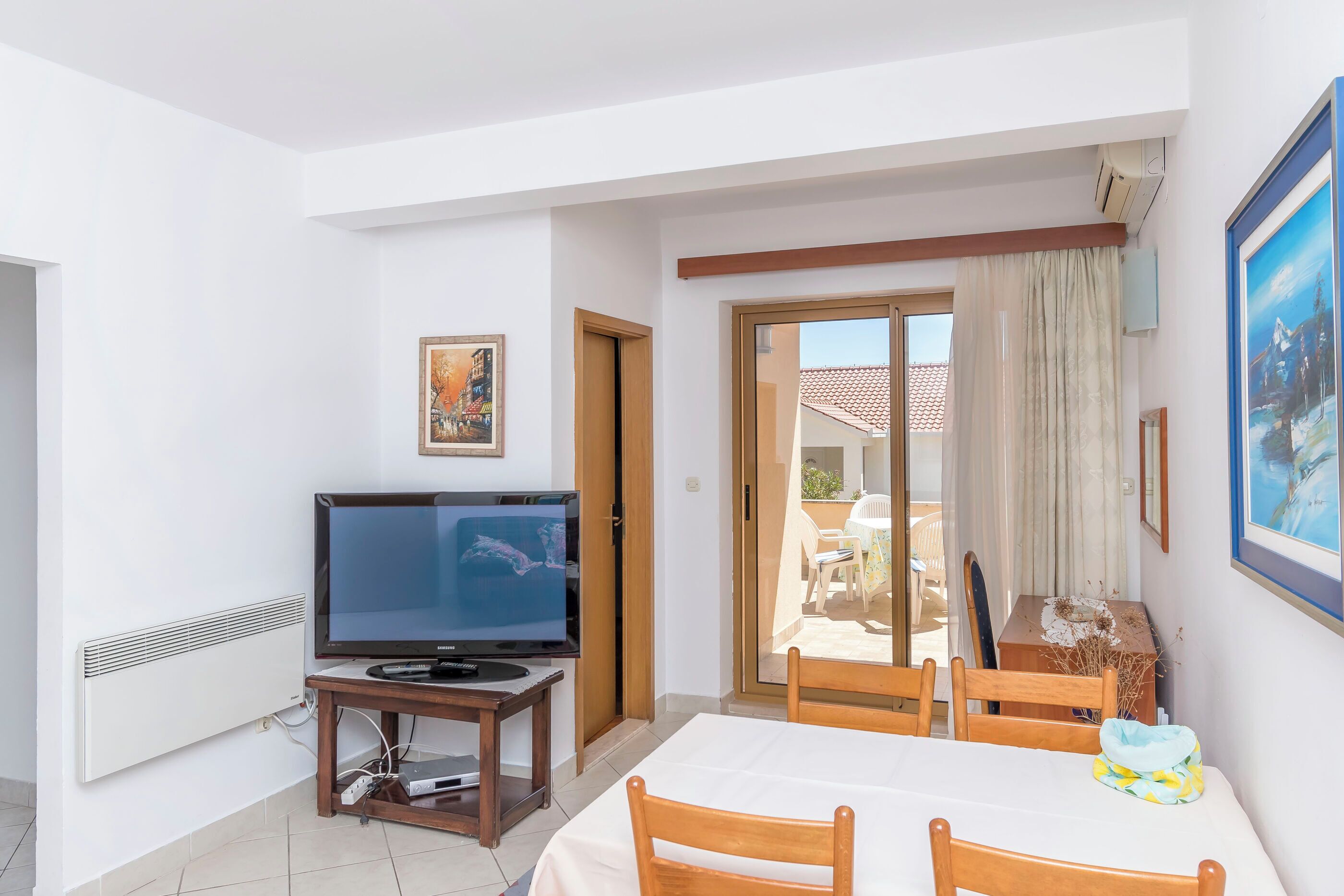 A room with a TV on a stand, a dining table with chairs, a door, and a view of the outside.