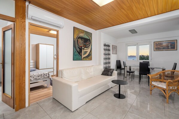 A modern living room with a white sofa, a dining table, and chairs. There is a painting on the wall and a window with a view.