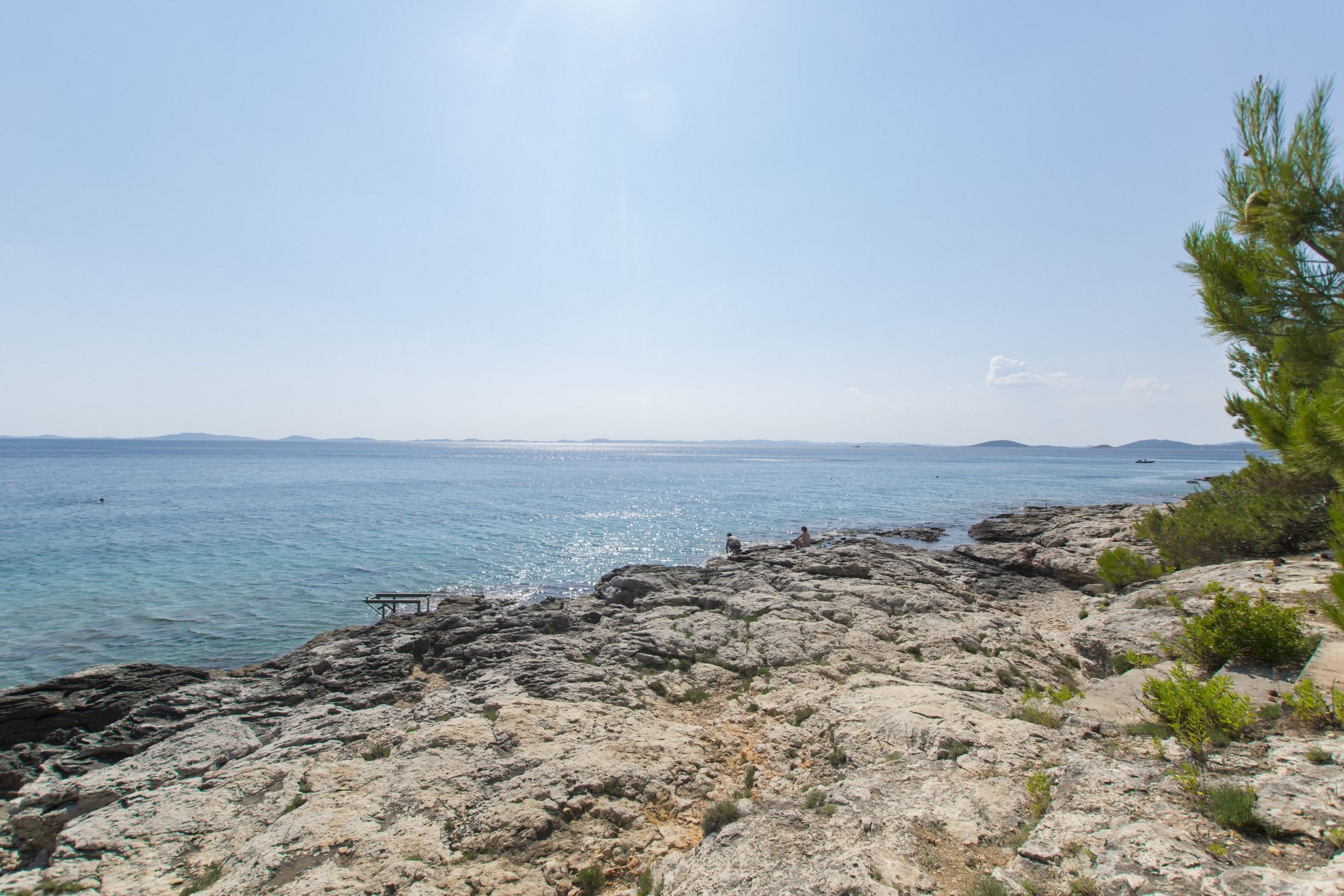 A rocky coastal landscape with clear blue sea, distant mountains, and sparse vegetation.
