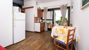 A compact kitchen with a refrigerator, microwave, and sink. A dining area with a table and chairs is visible, featuring a window with curtains. - Apartments Danica (Murter)