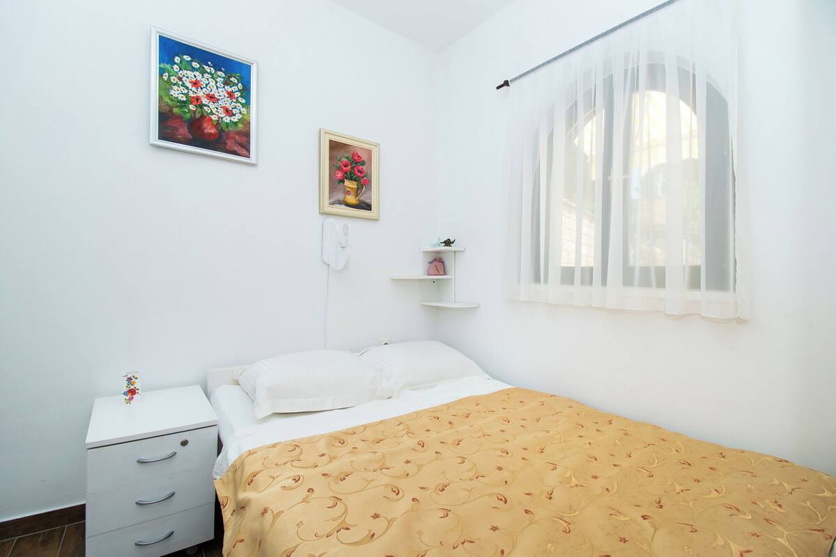 Une chambre à coucher comprenant un lit, une table de chevet, une commode, une fenêtre avec des rideaux légers et deux tableaux accrochés au mur.