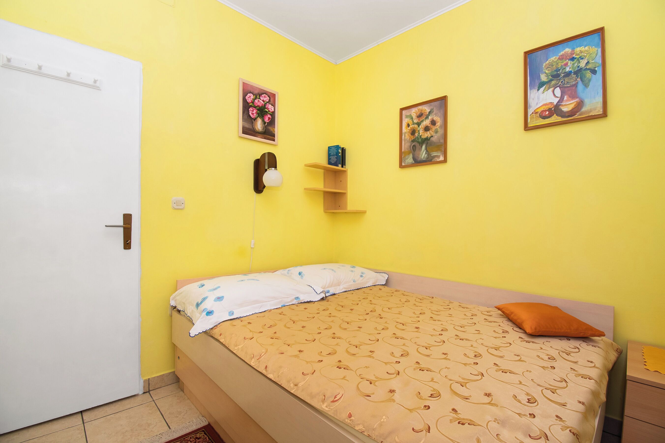Ein Zimmer mit einem Bett, einer weißen Tür, einer gelben Wand und zwei Bildern.