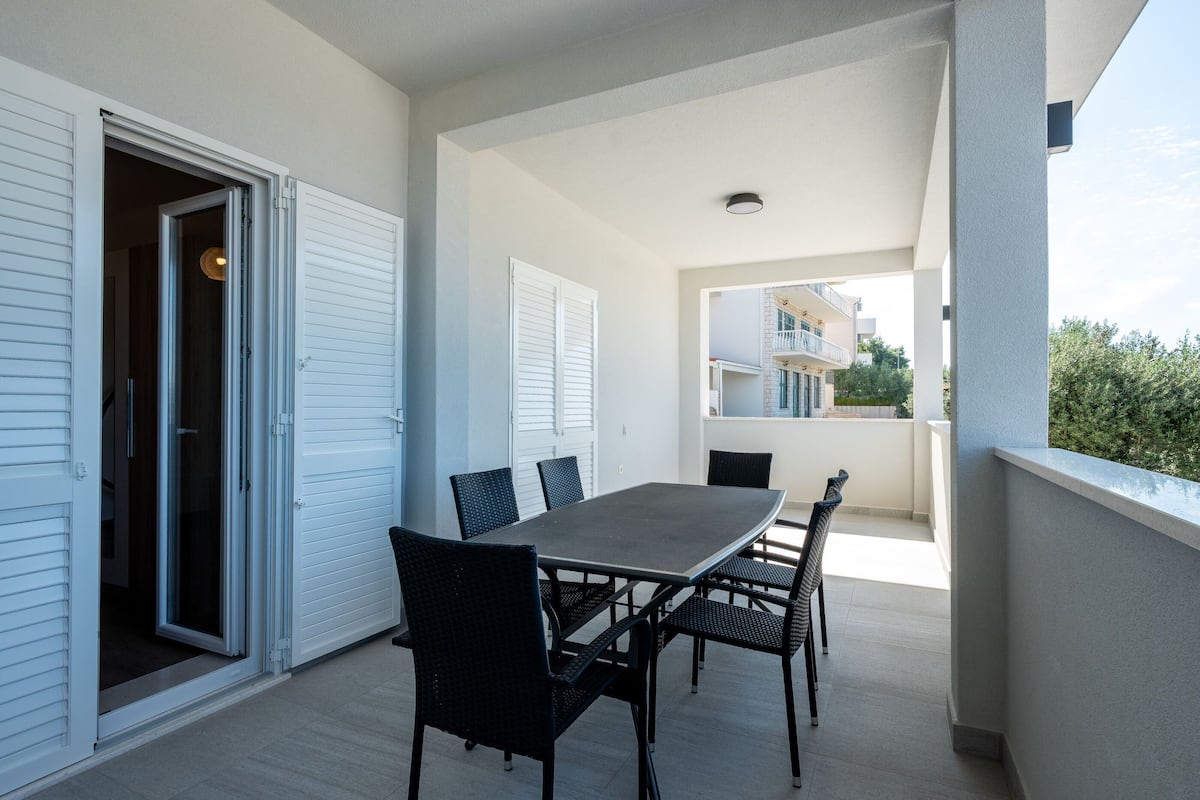 Un balcon extérieur avec une table et des chaises, une porte-fenêtre coulissante et une vue sur un bâtiment en arrière-plan.