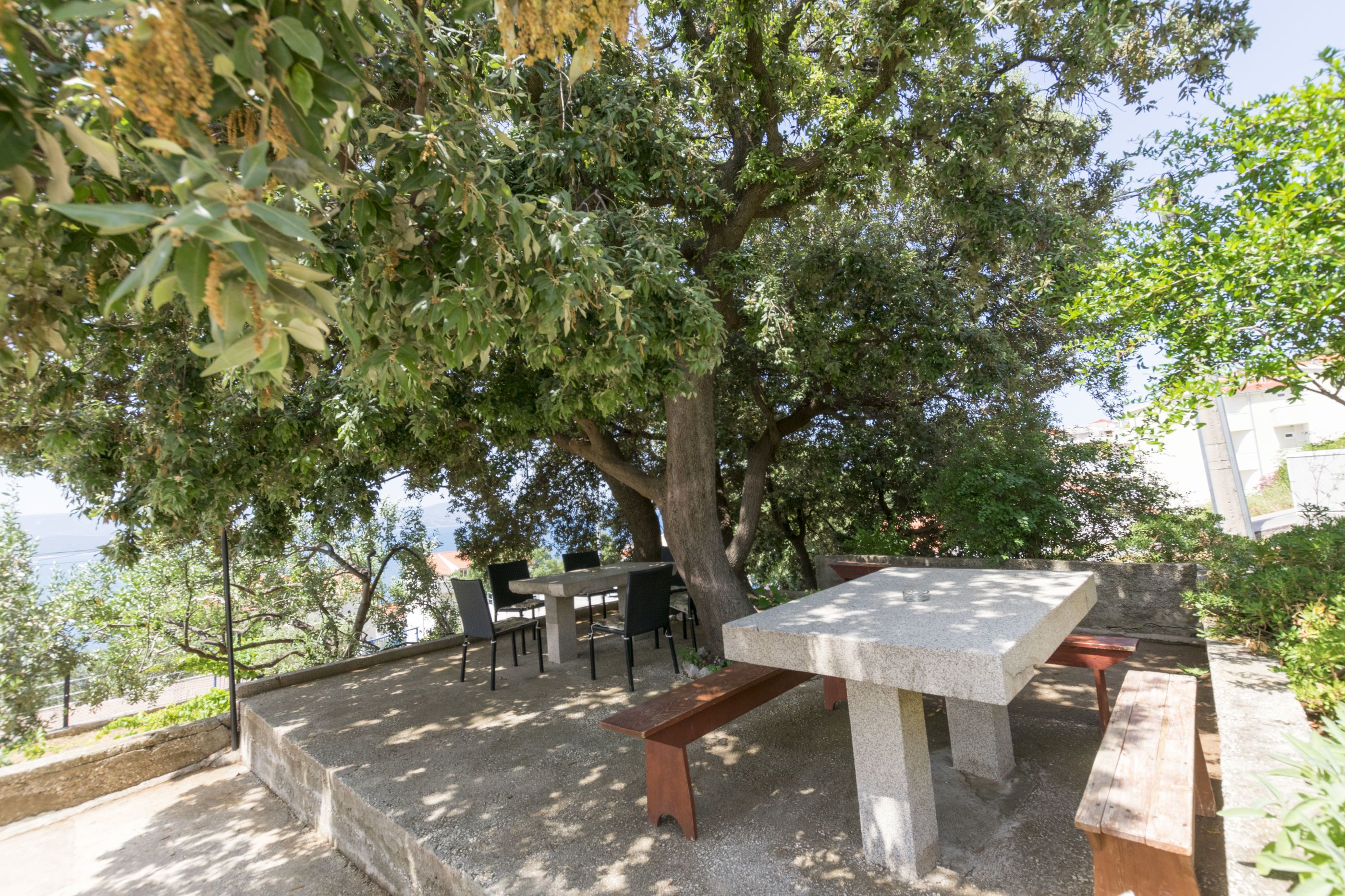 An outdoor dining area with a stone table and benches, surrounded by trees and a view of the sea.