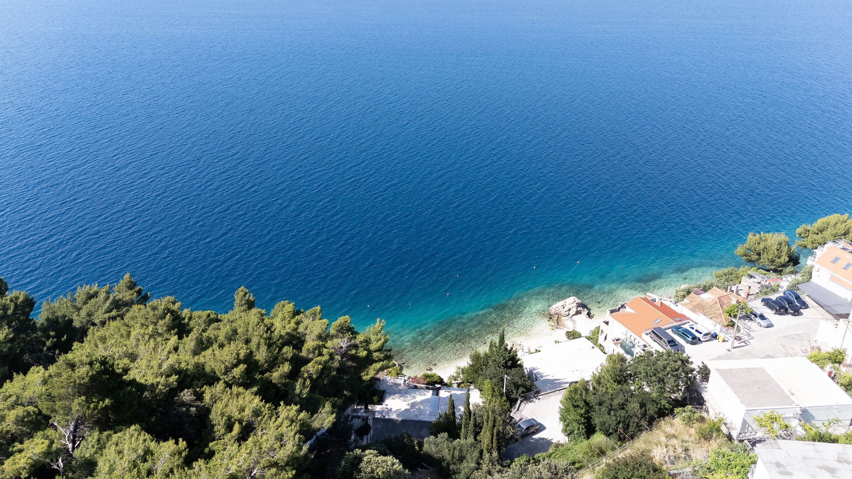 A coastal area with clear blue sea, a sandy beach, and buildings with parked cars.