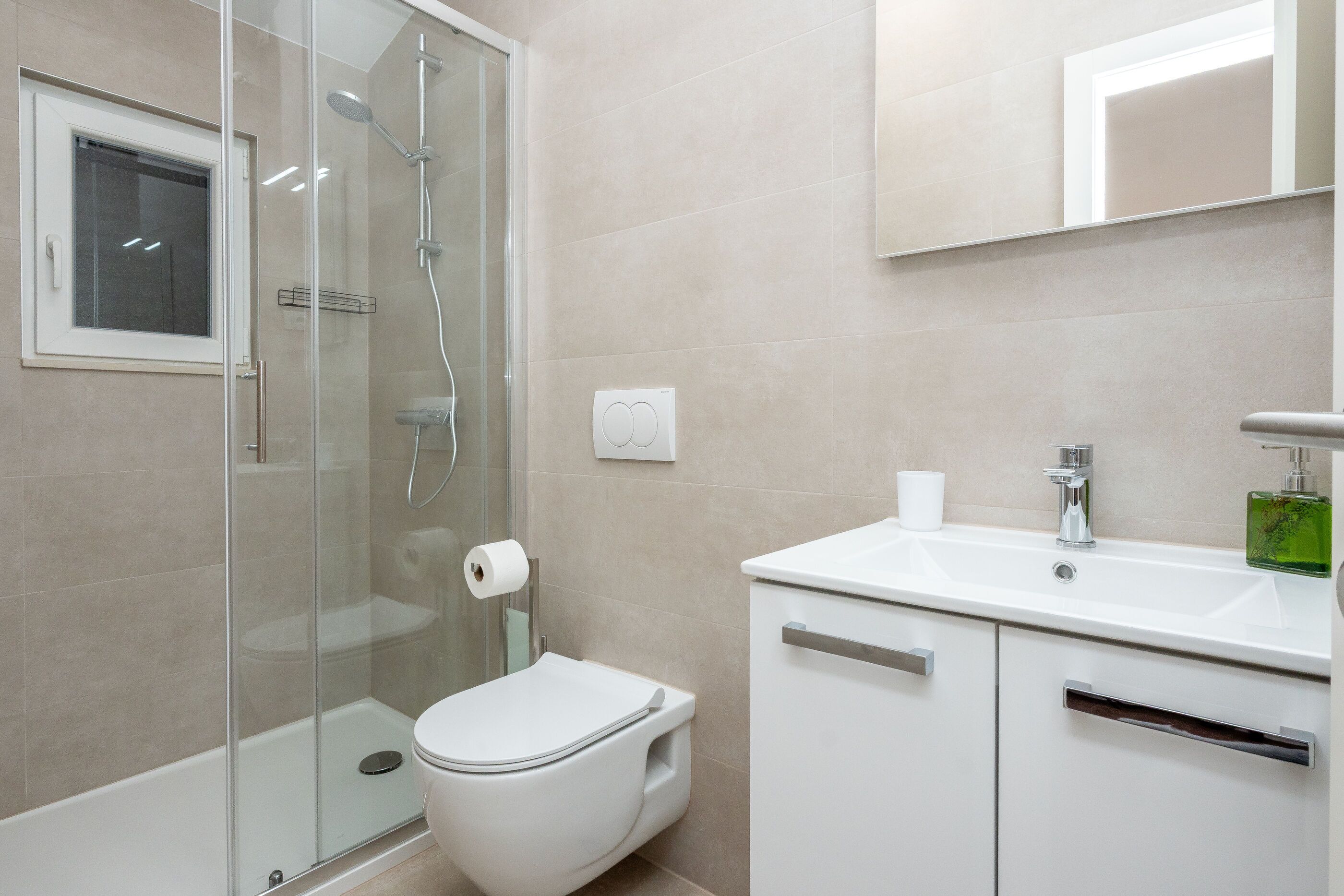 A modern bathroom with a glass shower enclosure, a toilet, a sink with a mirror above, and a green soap dispenser.