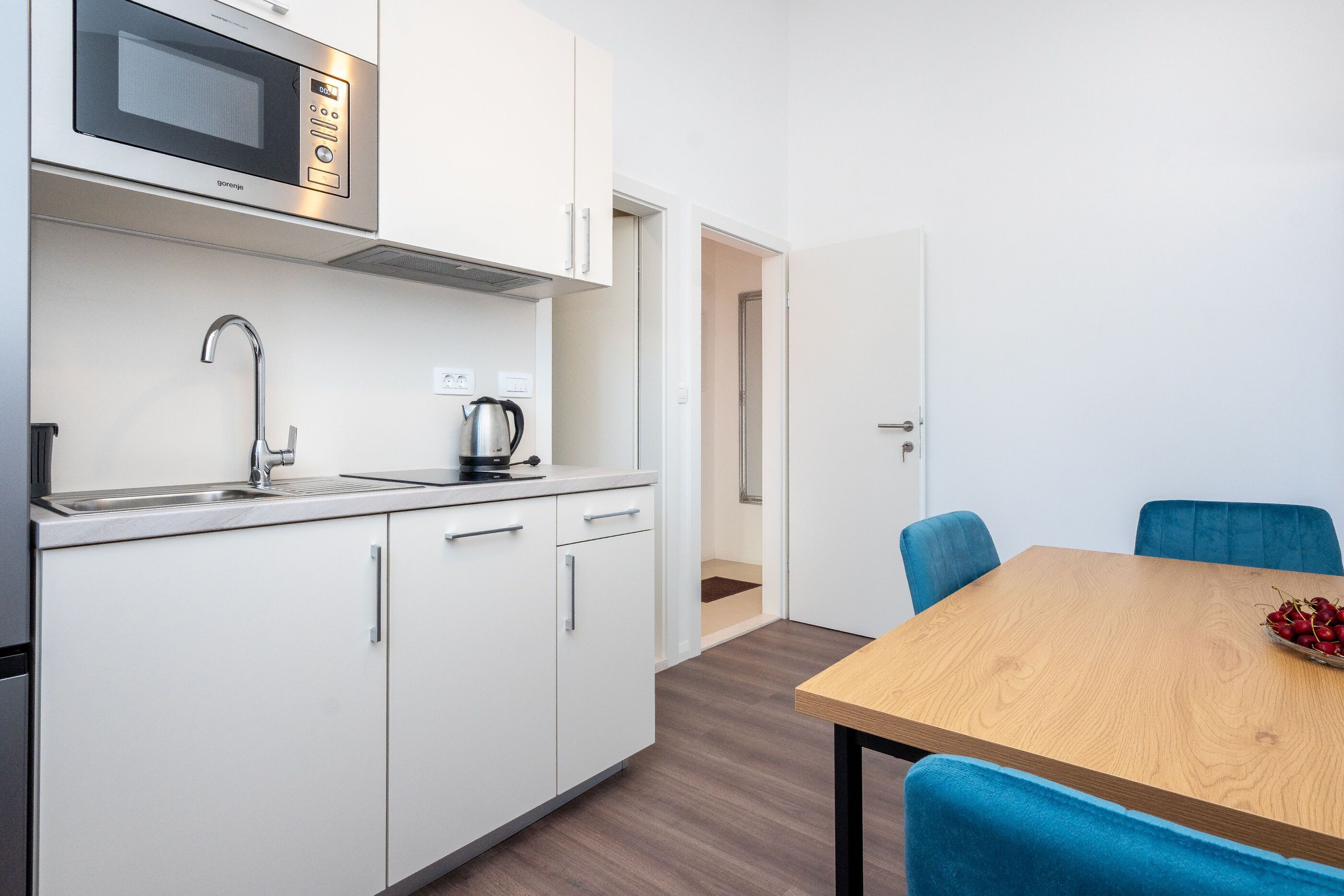 A modern kitchen with a microwave, sink, and kettle, a wooden dining table with blue chairs, and a door leading to another room.