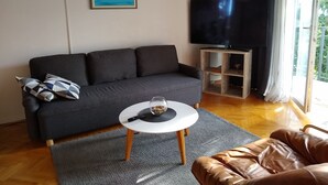 Uma sala de estar com um sofá cinza, uma mesa redonda pequena com um copo e uma televisão sobre um suporte de madeira.