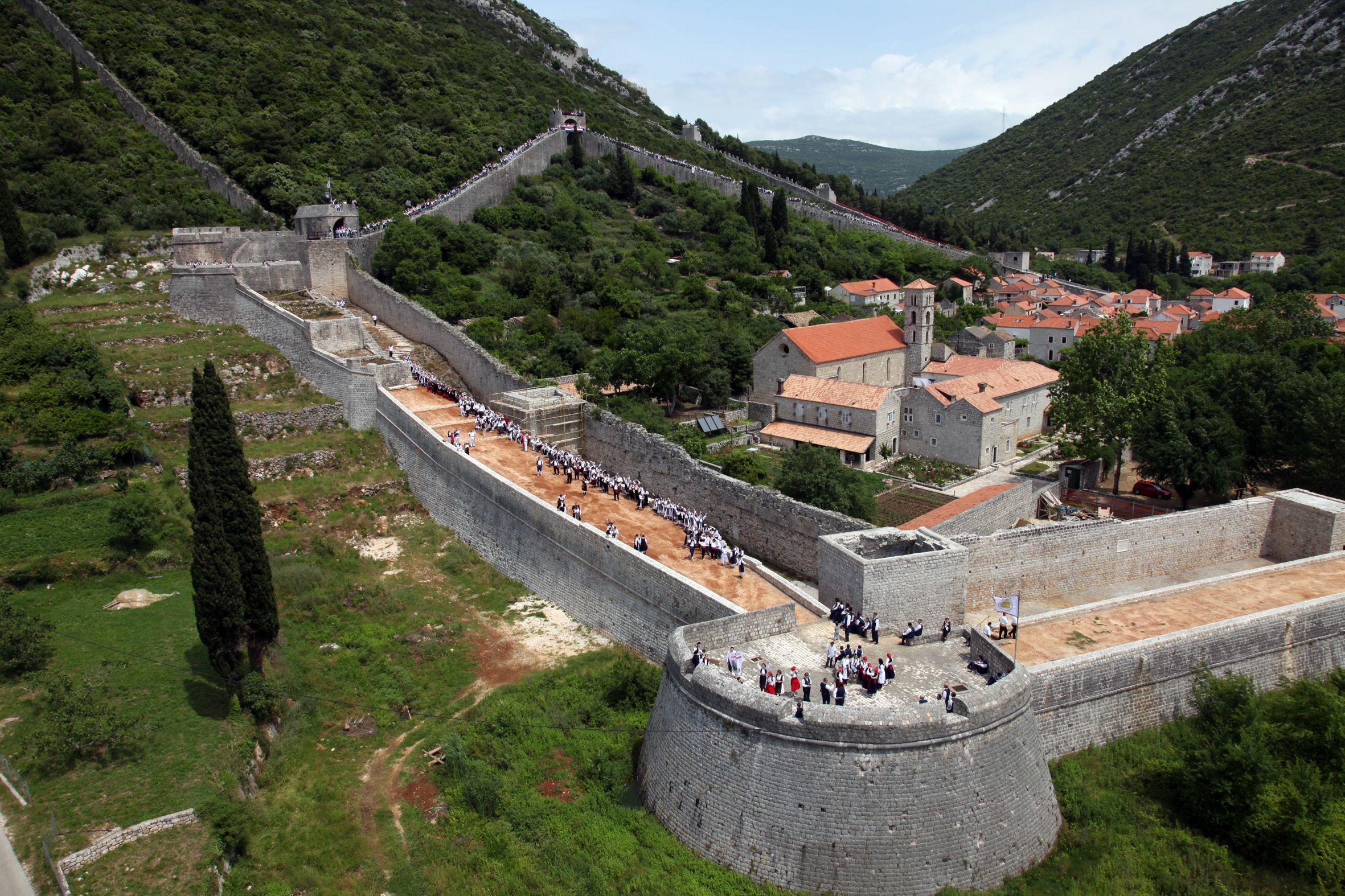 A historic stone wall with a crowd of people, surrounded by greenery and buildings with red roofs.
