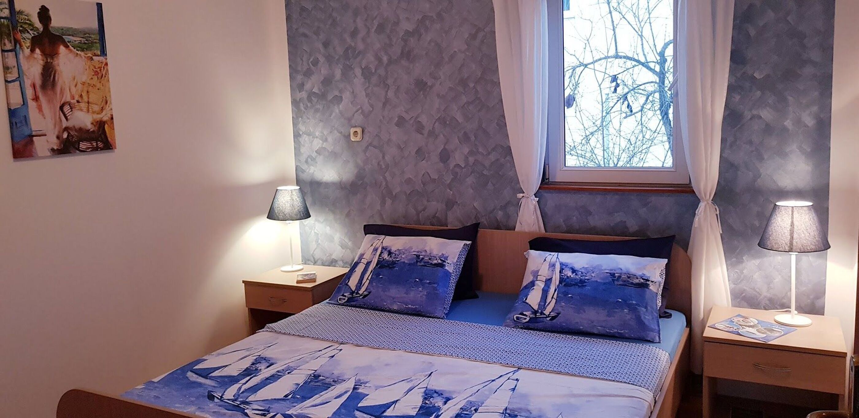 A bedroom with a bed, two nightstands, a window with curtains, and a wall-mounted painting.