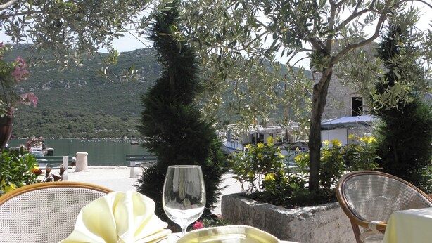 Une table dressée pour le dîner, avec un verre de vin blanc, une assiette et une serviette, donnant sur un plan d’eau et des collines.