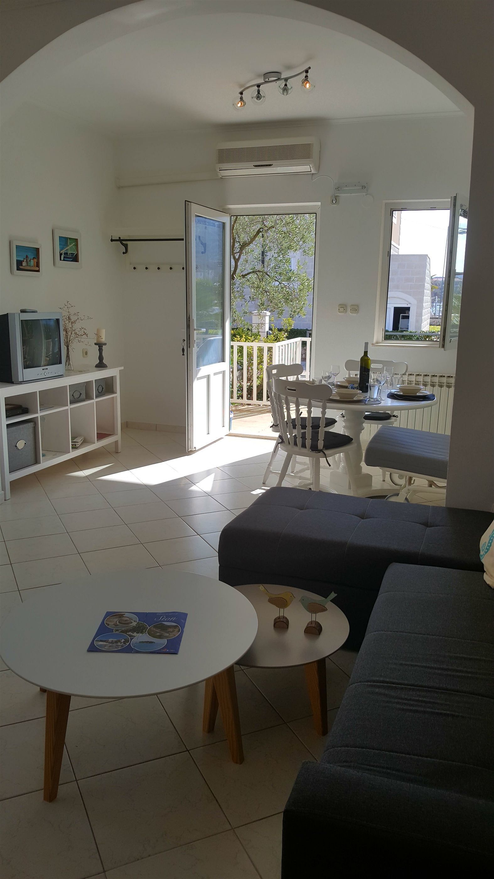 A living room with a sofa, a small table, a TV on a cabinet, and a balcony with a view of the outside.