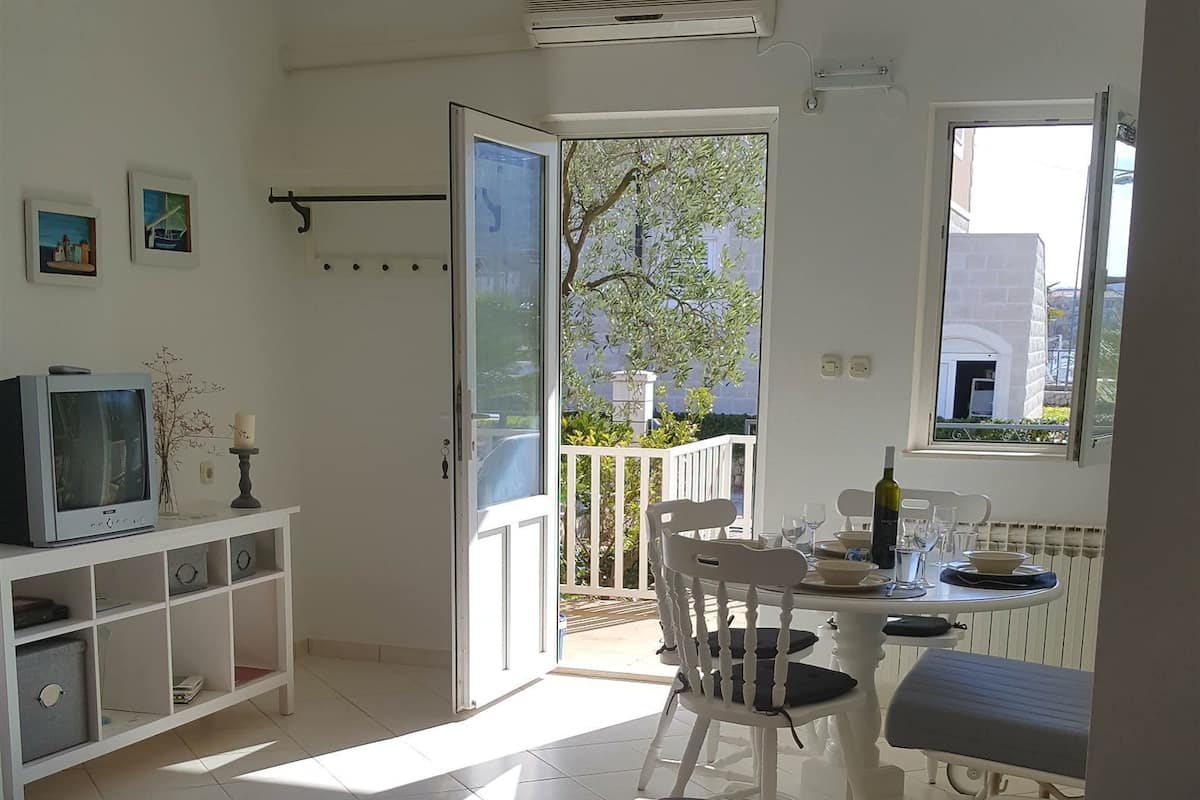 A living room with a sofa, a small table, a TV on a cabinet, and a balcony with a view of the outside.