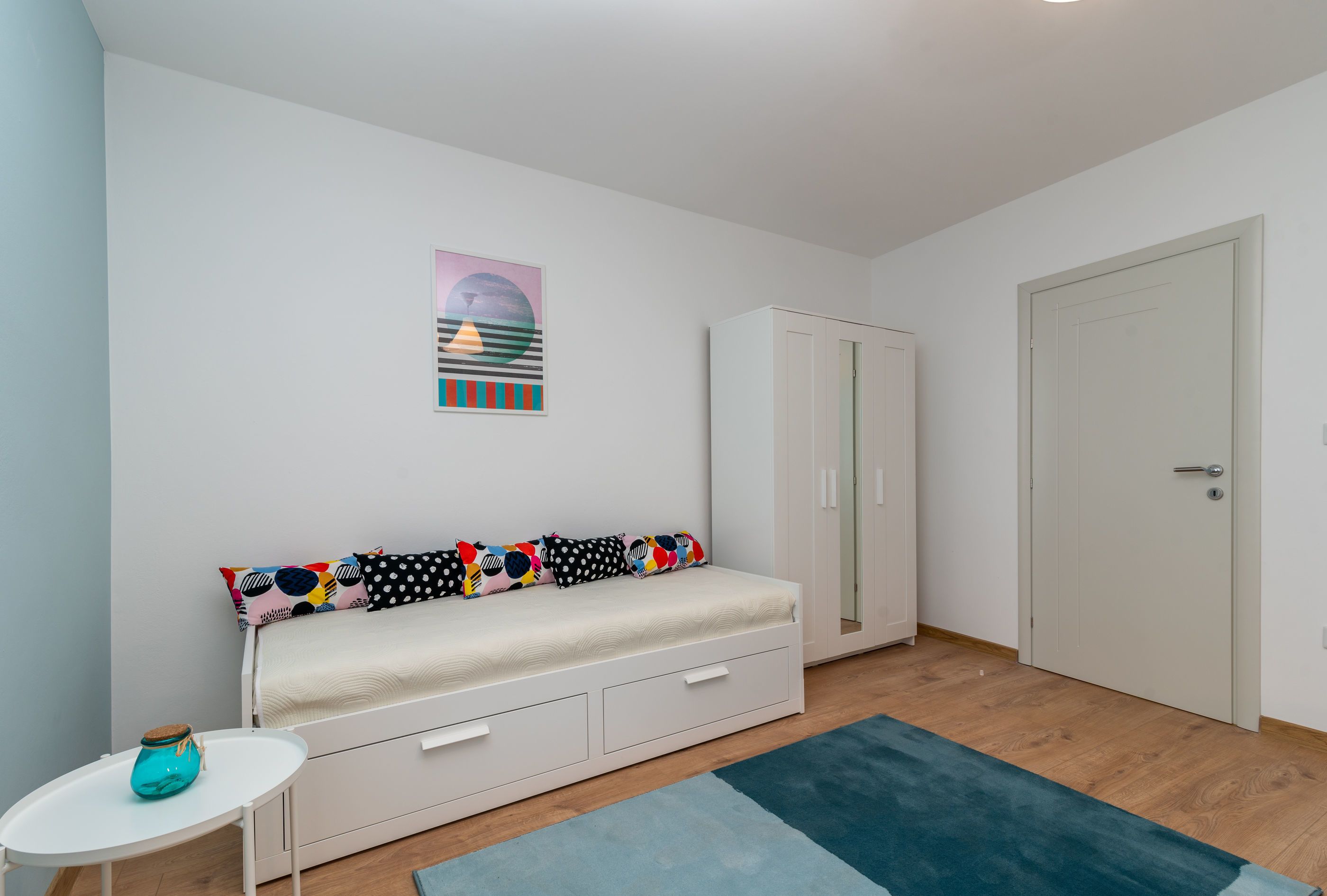 A room with a bed, a wardrobe, a small table with a vase, and a door.