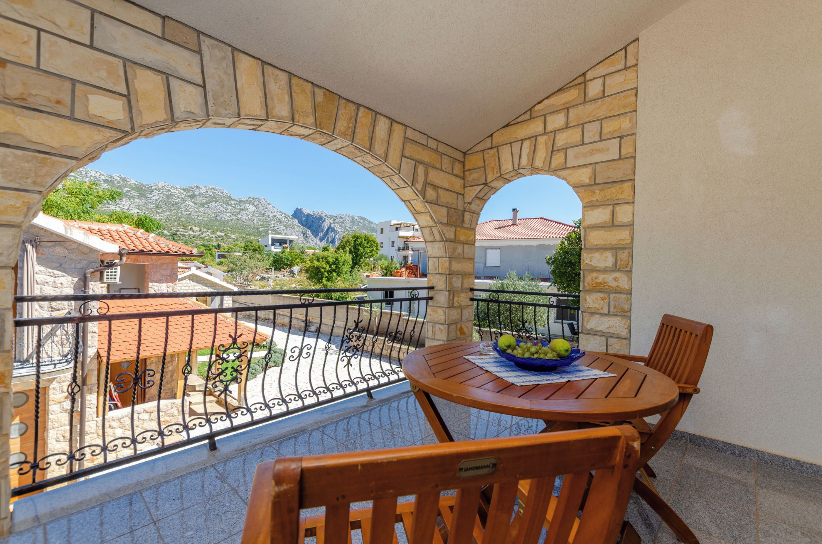 A balcony with stone arches, a table with a bowl of fruit, and a view of a mountainous landscape.