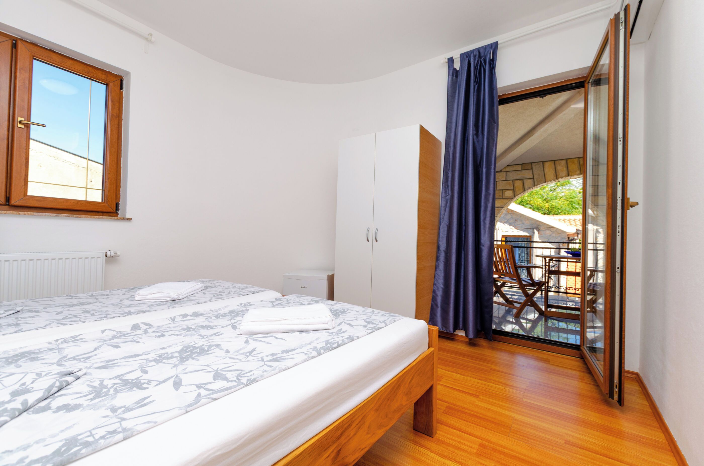 A bedroom with a bed, a wardrobe, a radiator, and a view of an outdoor area.