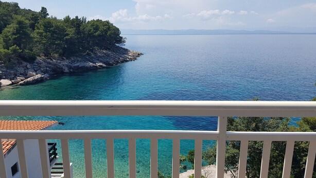 A balcony with a view of a clear blue sea and a rocky coastline with trees. - Apartments Teo i Ivana (Bogomolje)